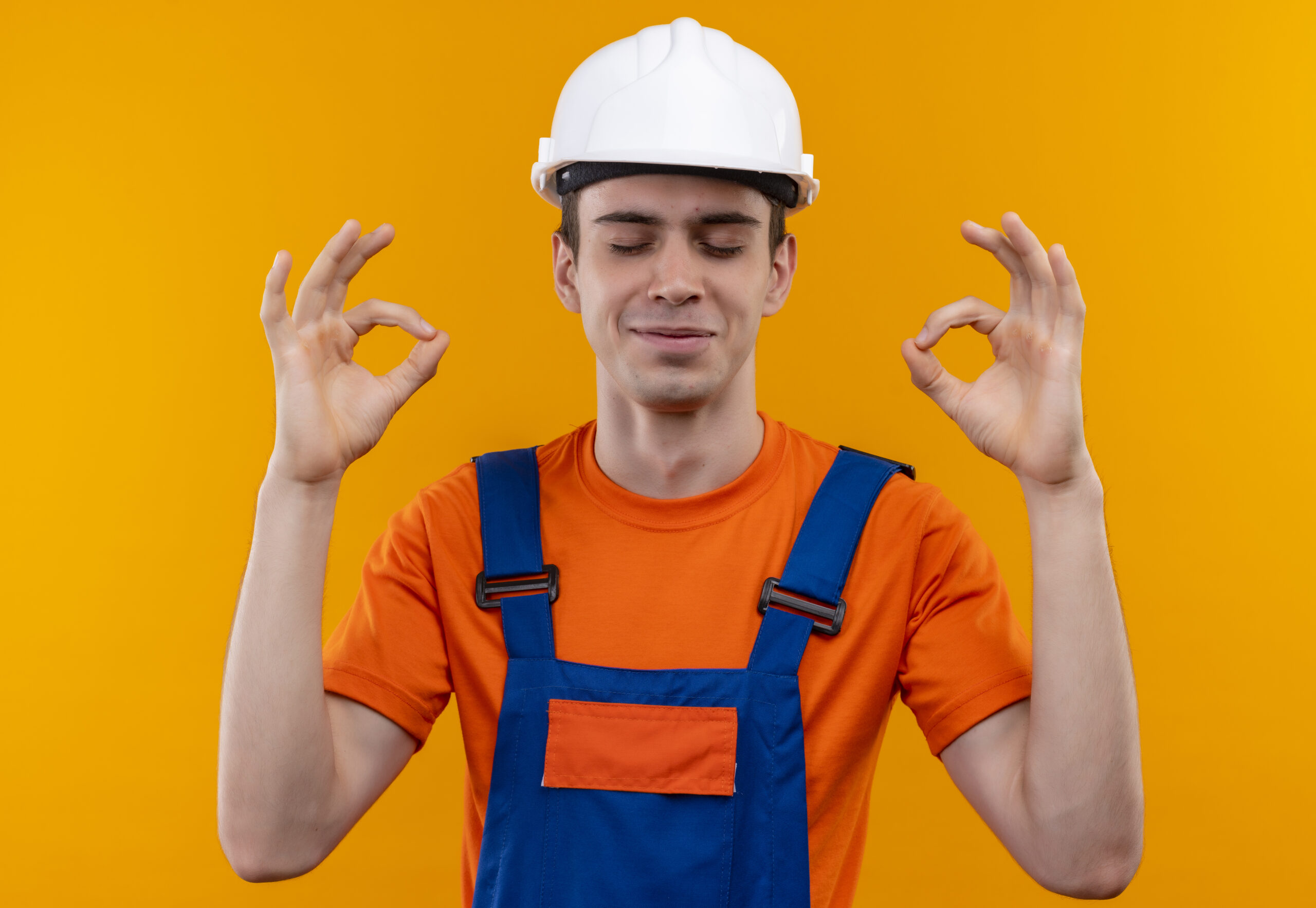 young builder man wearing construction uniform and safety helmet closing his eyes and doing ok with hands over orange isolated background